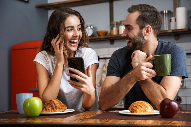 Beautiful cheerful couple having breakfast at the kitchen, using mobile phone