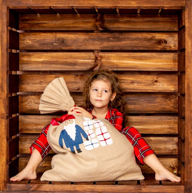 Beautiful cheerful girl in red Christmas pajamas holding a bag of gifts on a wooden background