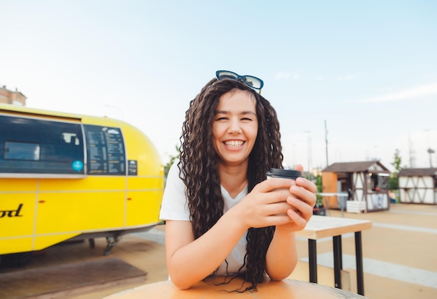 A beautiful cheerful girl with dreadlocks is sitting on a food\
court and drinking coffee fast food cafe on the street a woman\
drinks coffee in a cafe autumn