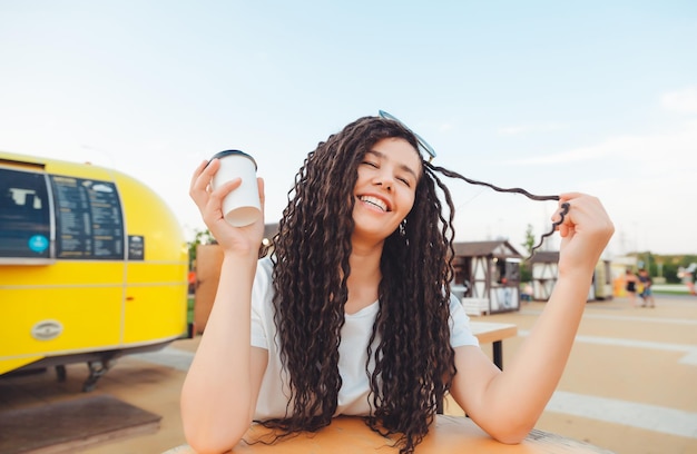 A beautiful cheerful girl with dreadlocks is sitting on a food court and drinking coffee fast food cafe on the street a woman drinks coffee in a cafe autumn