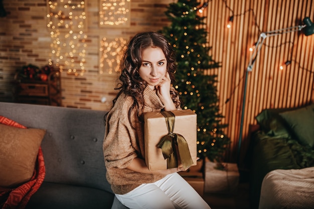 Beautiful cheerful happy young girl with christmas gifts on sofa on the background of a new year tree at home