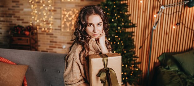 Beautiful cheerful happy young girl with christmas gifts on sofa on the background of a new year tree at home