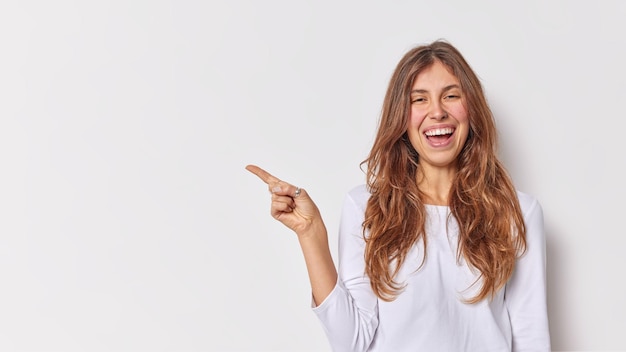 Beautiful cheerful long haired woman points index finger left shows logo banner smiles amused suggests to make shopping wears casual jumper isolated over white background demonstrates promo deal