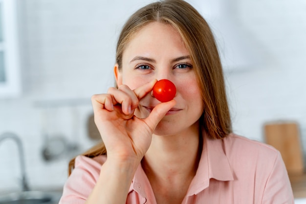 Beautiful cheerful person holds cherry tomato near nose. Ktchen blurred background. Funny facial expressions.