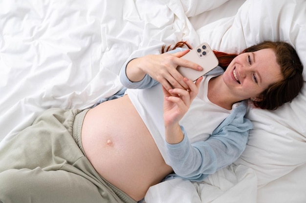 Beautiful cheerful pregnant woman taking selfie photo using mobile phone lying on bed at home big belly advanced pregnancy