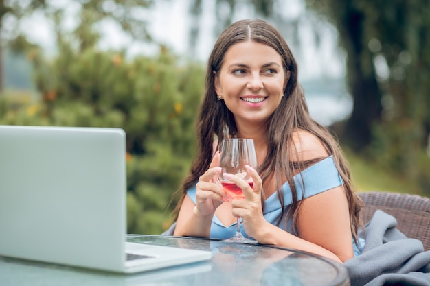 Bella donna allegra in vestito con un bicchiere di vino vicino al computer portatile in caffè estivo in uno stato d'animo sognante