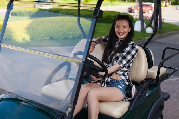 Beautiful cheerful woman with glasses on her head in   plaid shirt is sitting on a green golf cart.