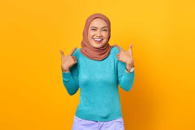 Beautiful cheerful young Asian woman pointing fingers herself on yellow background