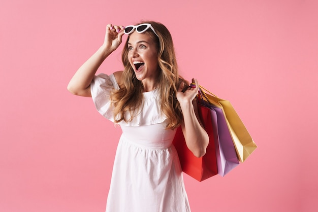 Beautiful cheerful young blonde girl wearing summer dress standing isolated over pink wall, carrying shopping bags