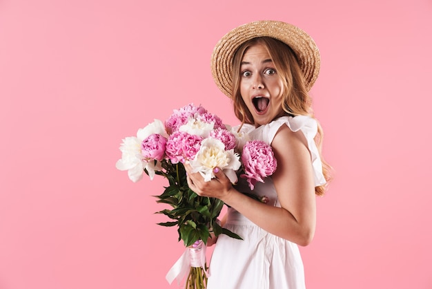 Beautiful cheerful young blonde girl wearing summer dress standing isolated over pink wall, holding bouquet of peonies
