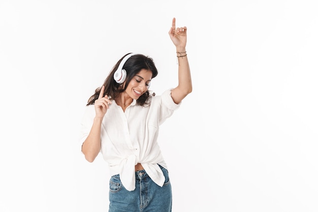 Bella giovane donna bruna allegra che indossa abiti casual in piedi isolata su bianco, ascoltando musica con cuffie wireless, ballando