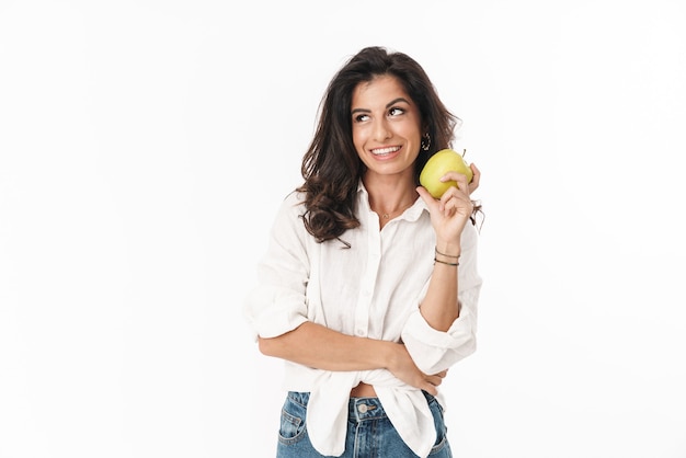 Beautiful cheerful young brunette woman wearing casual clothing standing isolated over white wall, showing green apple