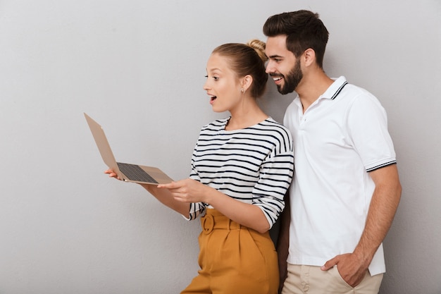 Beautiful cheerful young couple wearing casual clothing standing isolated over gray wall, using laptop computer