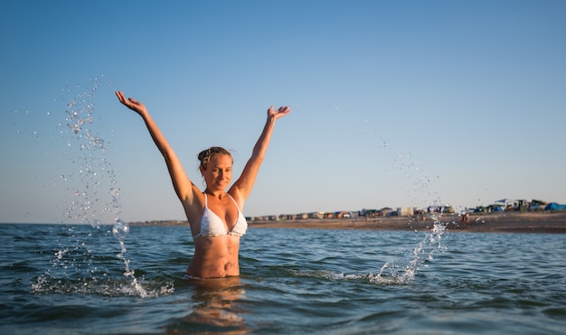 Beautiful cheerful young woman bathes in water and rejoices raising her hands up on sunny warm summer evening