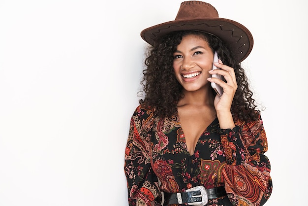 카우보이 모자를 쓰고 흰 벽에 격리된 채 휴대전화로 통화하는 아름다운 쾌활한 젊은 여성