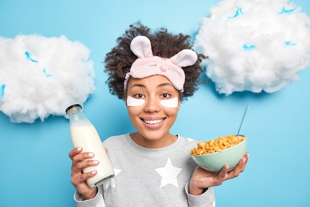 Beautiful cheerful young woman wears nightwear and sleepmask eats healthy food for breakfast holds cornflakes bowl and bottle of milk undergoes beauty procedures isolated over blue wall