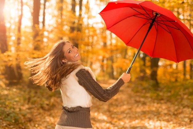 Beautiful cheerful young woman with red umbrella having fun in sunny forest in autumn colors.