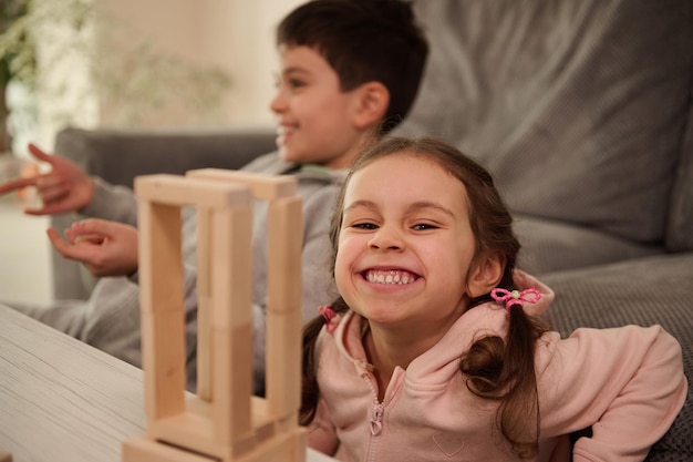 Photo beautiful child, adorable little girl in pink sweatshirt plays board game with her brother, builds wooden structures from wooden bricks and blocks, smiles toothy smile, looking at camera