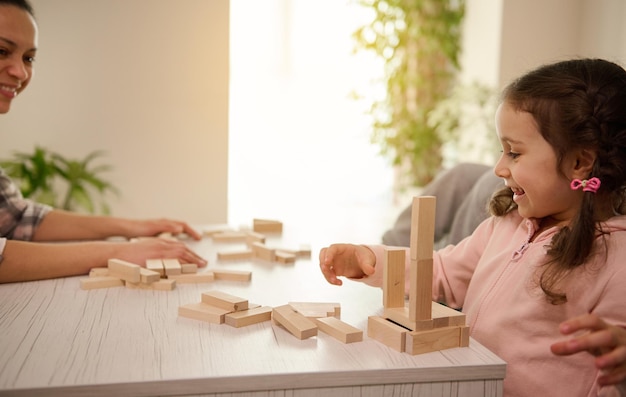 美しい子供、ピンクのスウェットシャツを着た愛らしい少女は、母親とボードゲームをし、木製のレンガとブロックから木製の構造物を作り、歯を見せる笑顔を浮かべ、教育的な娯楽を楽しんでいます