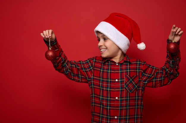 Beautiful child boy in checkered shirt and Santa Claus hat looking at Christmas balls toys in his hands, isolated over red colored background with copy space for advertisement