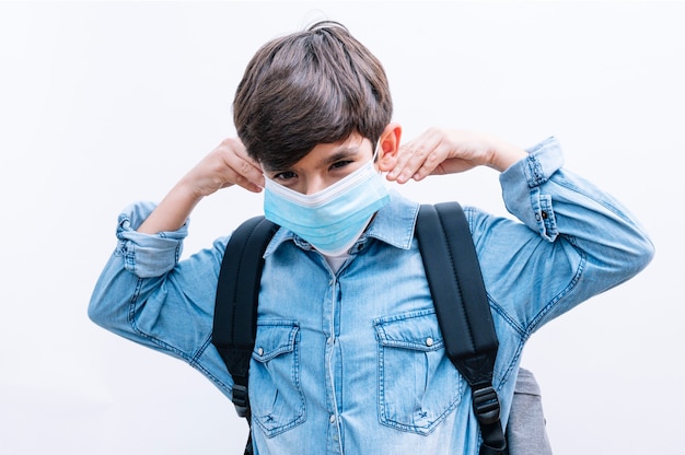 Bello studente del ragazzo del bambino con lo zaino e la maschera che tengono i libri su fondo bianco