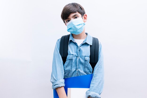 Beautiful child boy student with backpack and mask holding books on white background