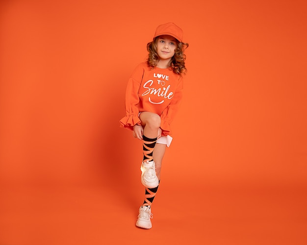 Красивая детская девочка 6-7 лет в кепке в кроссовках прыгает и танцует на оранжевом фоне. Рекламное фото для танцевальной студии с копией пространства