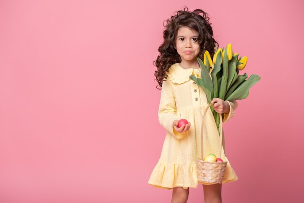 Beautiful child girl holding basket with easter eggs and tulips on pink background.