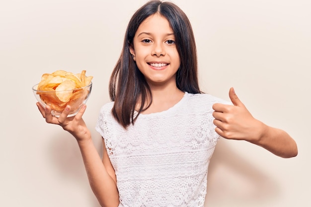 Beautiful child girl holding potato chip smiling happy and positive thumb up doing excellent and approval sign