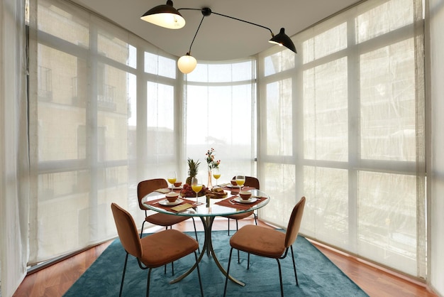 Beautiful circular beveled edge clear glass dining table with crockery of fresh fruit and sweet desserts brown velvet upholstered chairs and bay window with blinds