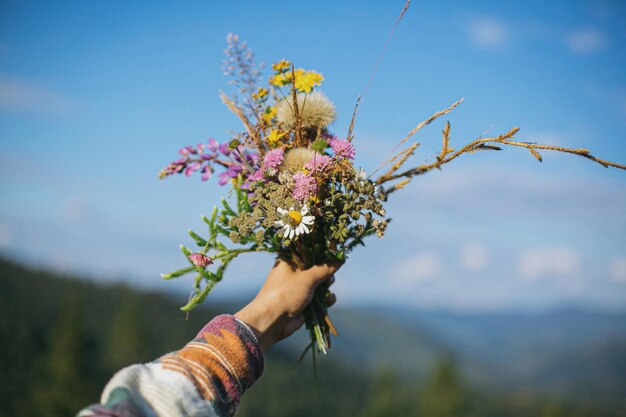 Foto bellissimo bouquet colorato di fiori di campo in mano sullo sfondo di colline di montagna e cielo soleggiato