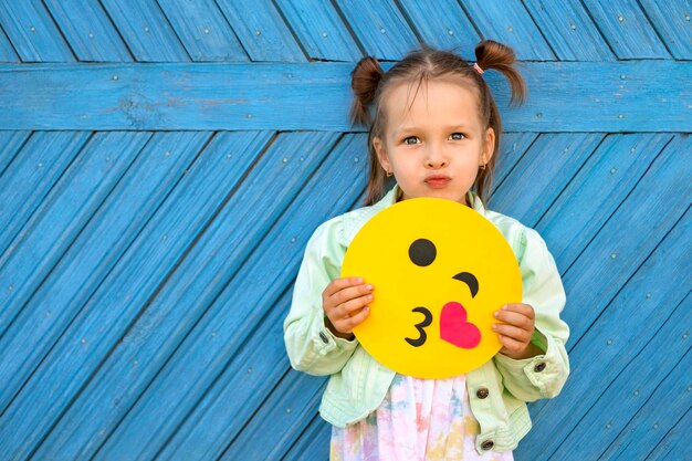 Beautiful comical girl sends an air kiss holding yellow funny emoticon in hand