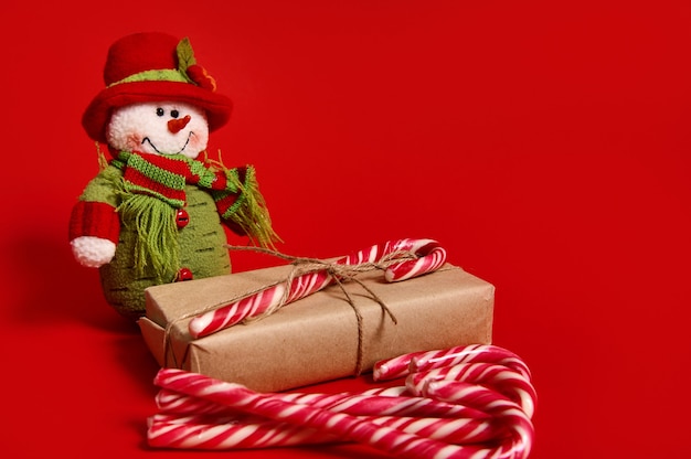 크리스마스 품목의 아름다운 구성, 묶인 활이 있는 크래프트 포장지에 있는 선물 상자, 광고용 복사 공간이 있는 빨간색 배경 위에 격리된 달콤한 사탕 지팡이 및 눈사람 봉제 장난감