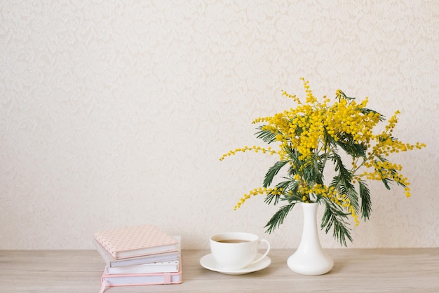 Beautiful composition of mimosa branches and a white tea cup with a saucer and a stack of notebooks on a table with a copy space