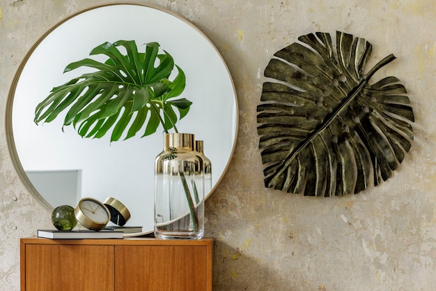 Beautiful composition at modern retro interior with design\
commode, tropical leaf in vase, round mirror, clock, book, grunge\
wall and elegant accessories in stylish home decor.