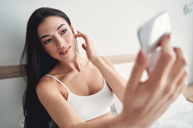 Beautiful concentrated Caucasian young woman in a bra photographing herself with a smartphone in bed