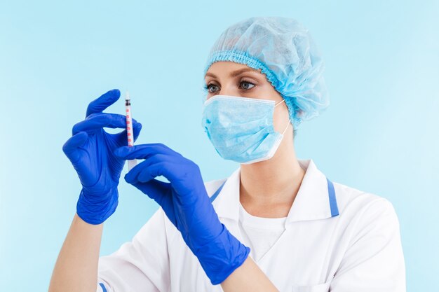 Beautiful confident blonde woman doctor wearing uniform and mask standing isolated over blue wall, holding syringe