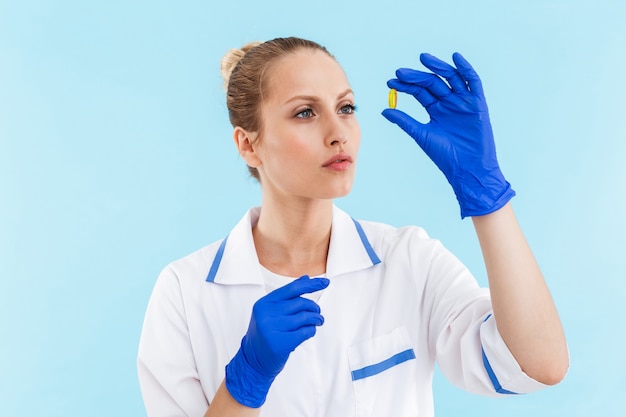 아름 다운 자신감 금발 여자 의사는 약을 보고 파란색 벽에 고립 된 제복을 입고 서