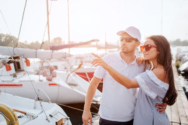 Beautiful and confident couple stands on pier and hug each other. They stand very close. Girl point with finger