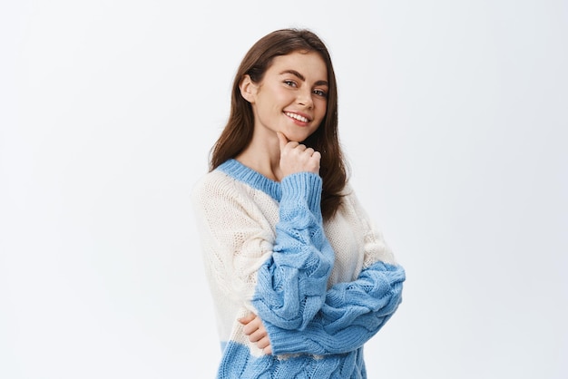 Beautiful confident girl holding hand on chin and smiling at camera with coquettish look standing in sweater thinking with pensive face expression white background