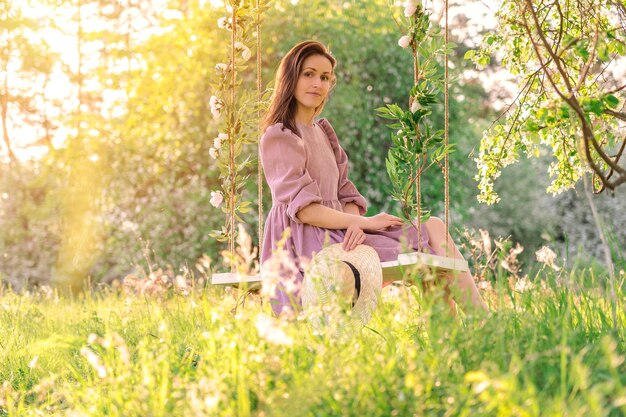 A beautiful confident girl in a purple dress poses on a rope swing in a blooming apple orchard