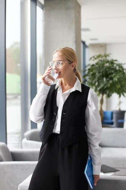 현대 사무실에서 창문을 바라보며 물을 마시는 아름다운 자신감 있는 여성