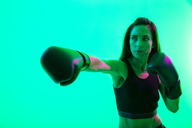 Beautiful confident young fitness girl standing isolated over green neon wall, boxing