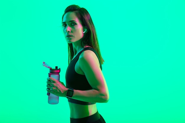 Beautiful confident young fitness girl standing isolated over green neon wall, wearing wireless earphones, holding water bottle after workout