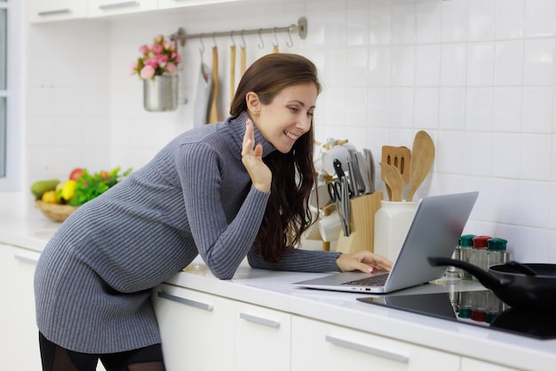 Beautiful cool pregnant woman standing in modern kitchen and using laptop computer to chat in videocall with happy and relaxing manner