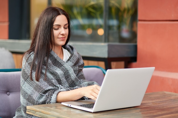 Beautiful copywriter keyboards on laptop computer, provides marketing service online, wrapped in coverlet, poses in cozy coffee shop