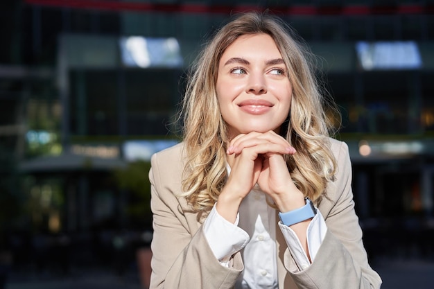 Beautiful corporate woman looks dreamy and smiles stands outside on street leans her head on hands d