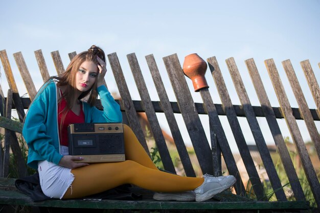 A beautiful country girl in bright clothes sits on a wooden bench with a cassette recorder