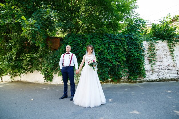 Bella coppia sposa in un abito bianco con un bouquet mentre lo sposo con bretelle e papillon si tengono per mano. sullo sfondo di un muro con foglie verdi. coppia felice. concetto di matrimonio.