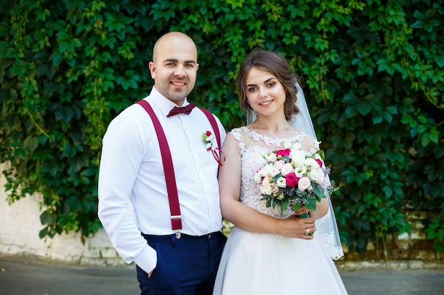 Beautiful couple bride in a white dress with a bouquet while the groom with suspenders and bow tie holding hands. Against the background of a wall with green leaves. Happy couple. Marriage concept.